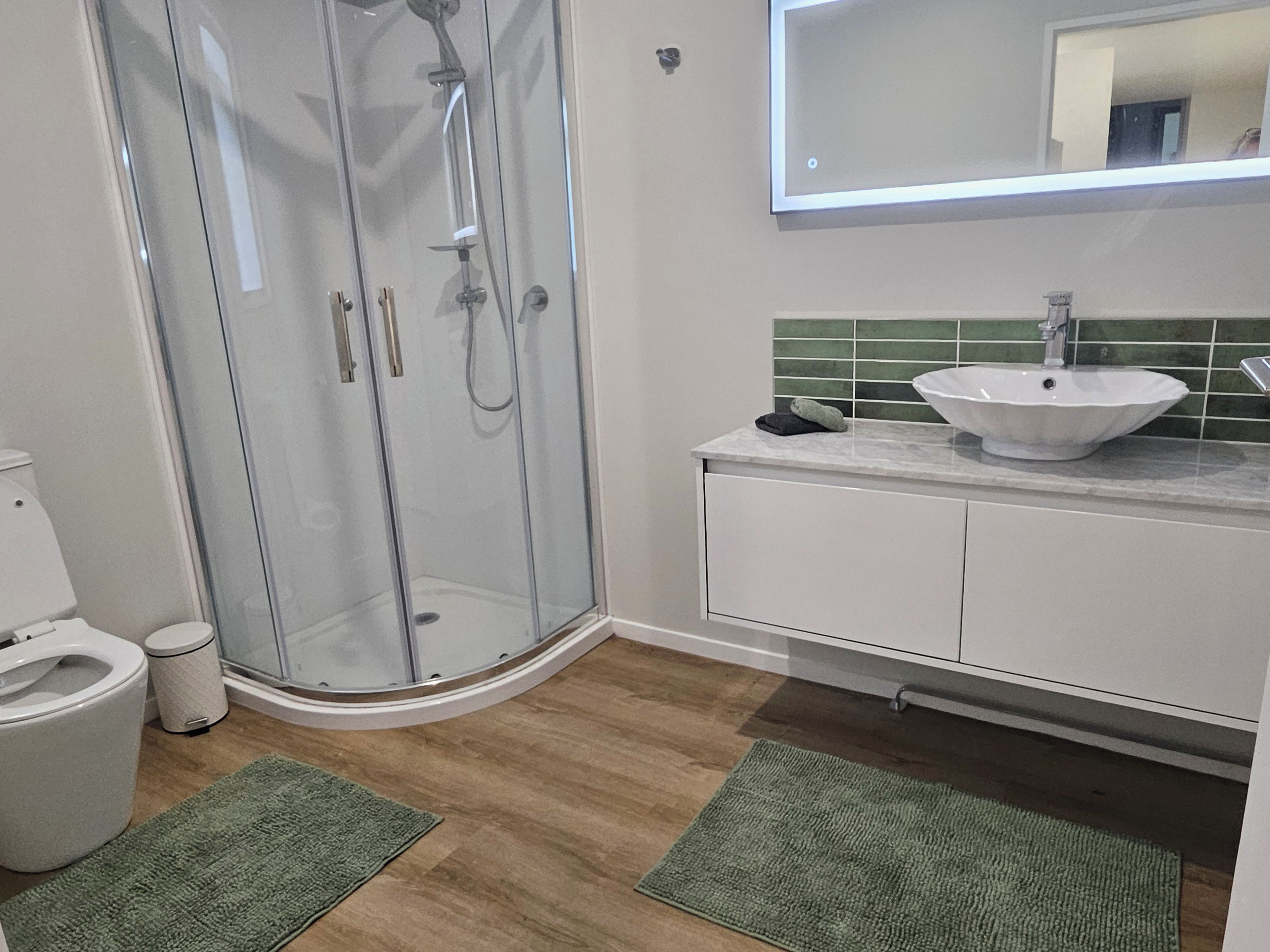 A bathroom with a glass-enclosed shower, a white sink, and a green-tiled backsplash.