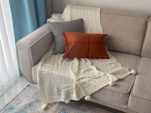 En sofa med et hvidt strikket tæppe, to puder (en med mønster, en ensfarvet) og en lille måtte med kvaster.