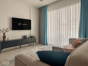 A modern living room with a flat-screen TV mounted on the wall, a dark-colored console table, a sofa with a blue accent, and sheer curtains on the window.