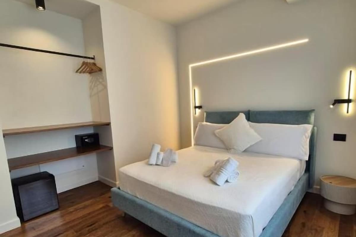 A modern hotel room with a large bed, a bedside table, a wardrobe, and a small seating area.