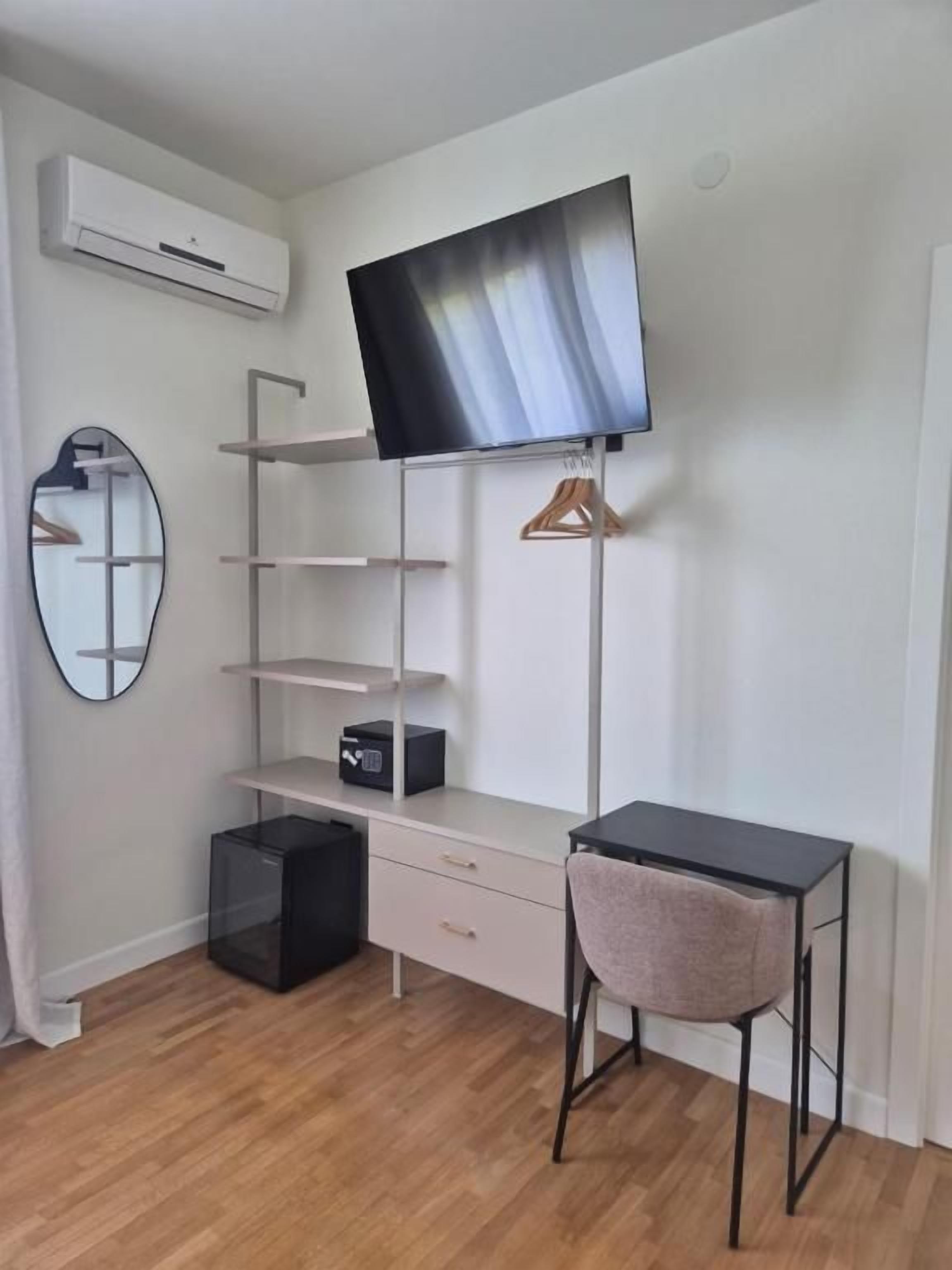 A modern room with a flat-screen TV mounted on the wall, a desk with a chair, a mirror, and a small black storage unit.