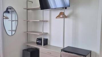 A modern room with a flat-screen TV mounted on the wall, a desk with a chair, a mirror, and a small black storage unit.
