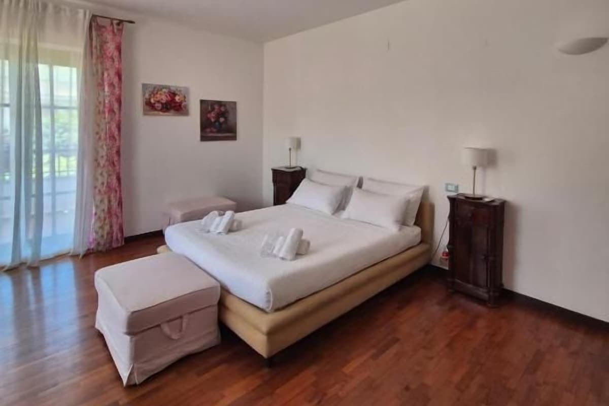 A bedroom with a bed, bedside tables, a nightstand, a window with curtains, and a wooden floor.