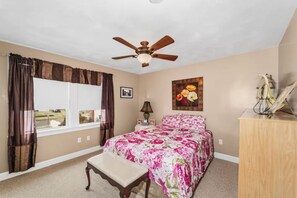 A bedroom with a bed, a ceiling fan, a window with curtains, a bedside table, a lamp, and a painting. - The Salty Escape (Narragansett)