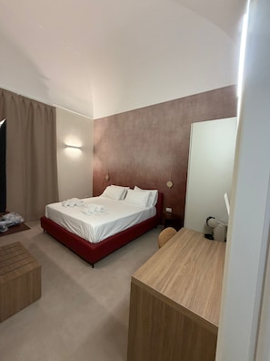 A hotel room with a bed, a desk, a chair, a mirror, and a wall-mounted light fixture. - Alkimia Luxury Rooms (BRINDISI)