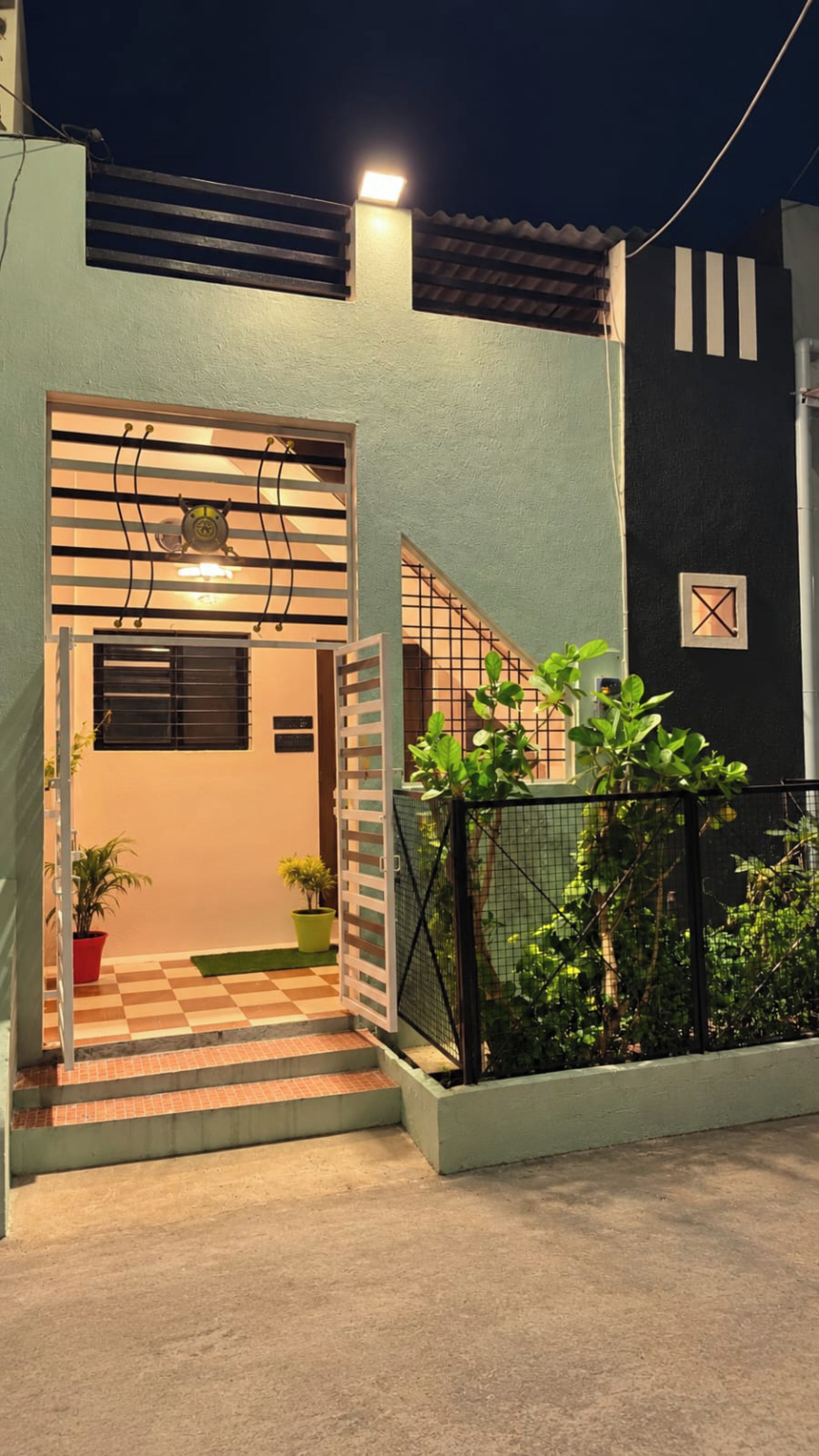 A residential building with a gated entrance, a staircase leading to the entrance, and a small garden area with potted plants.