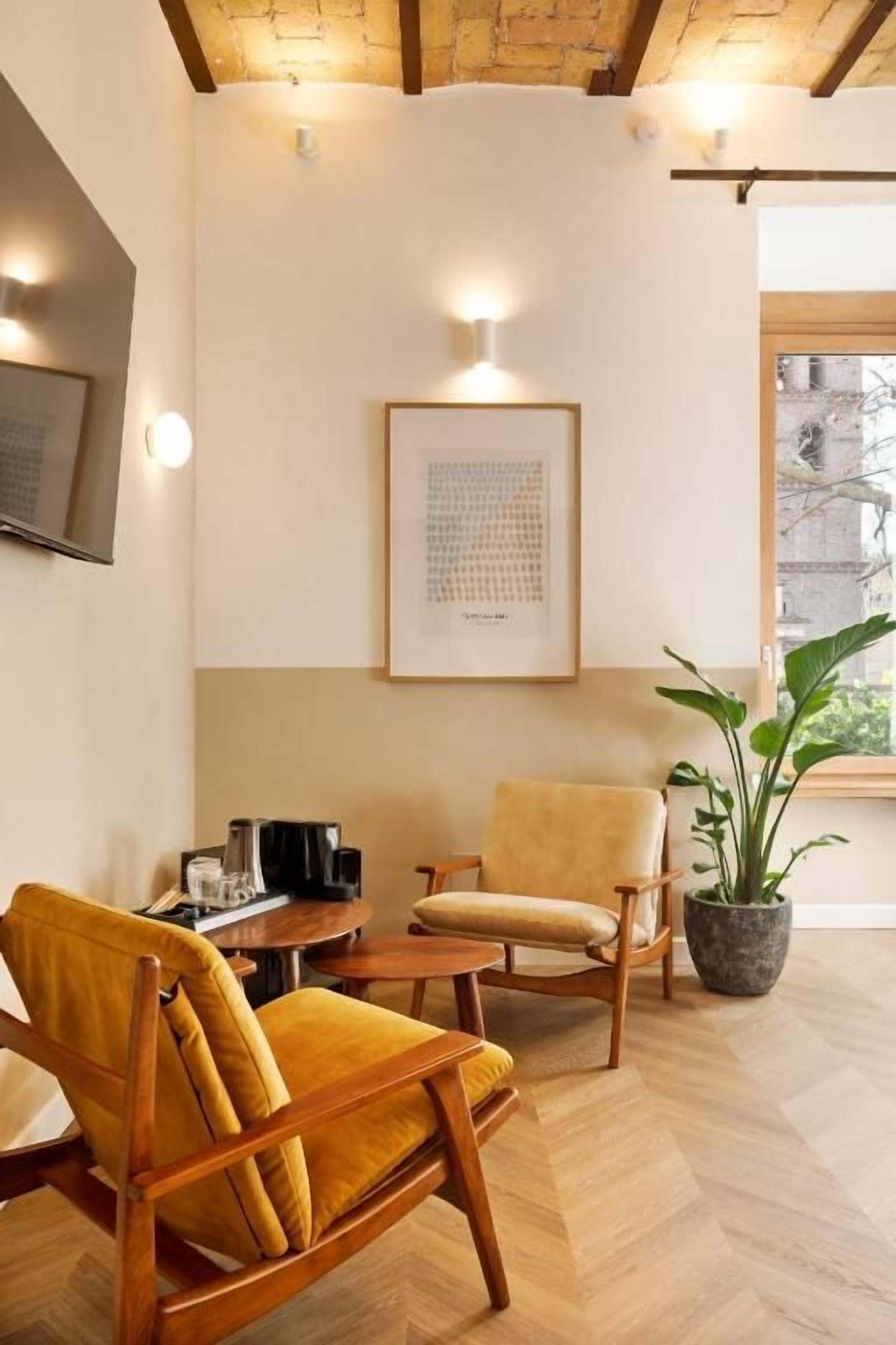 Una habitación con piso de espiga, dos sillas de madera, una mesita, una planta en maceta y un cuadro en la pared.