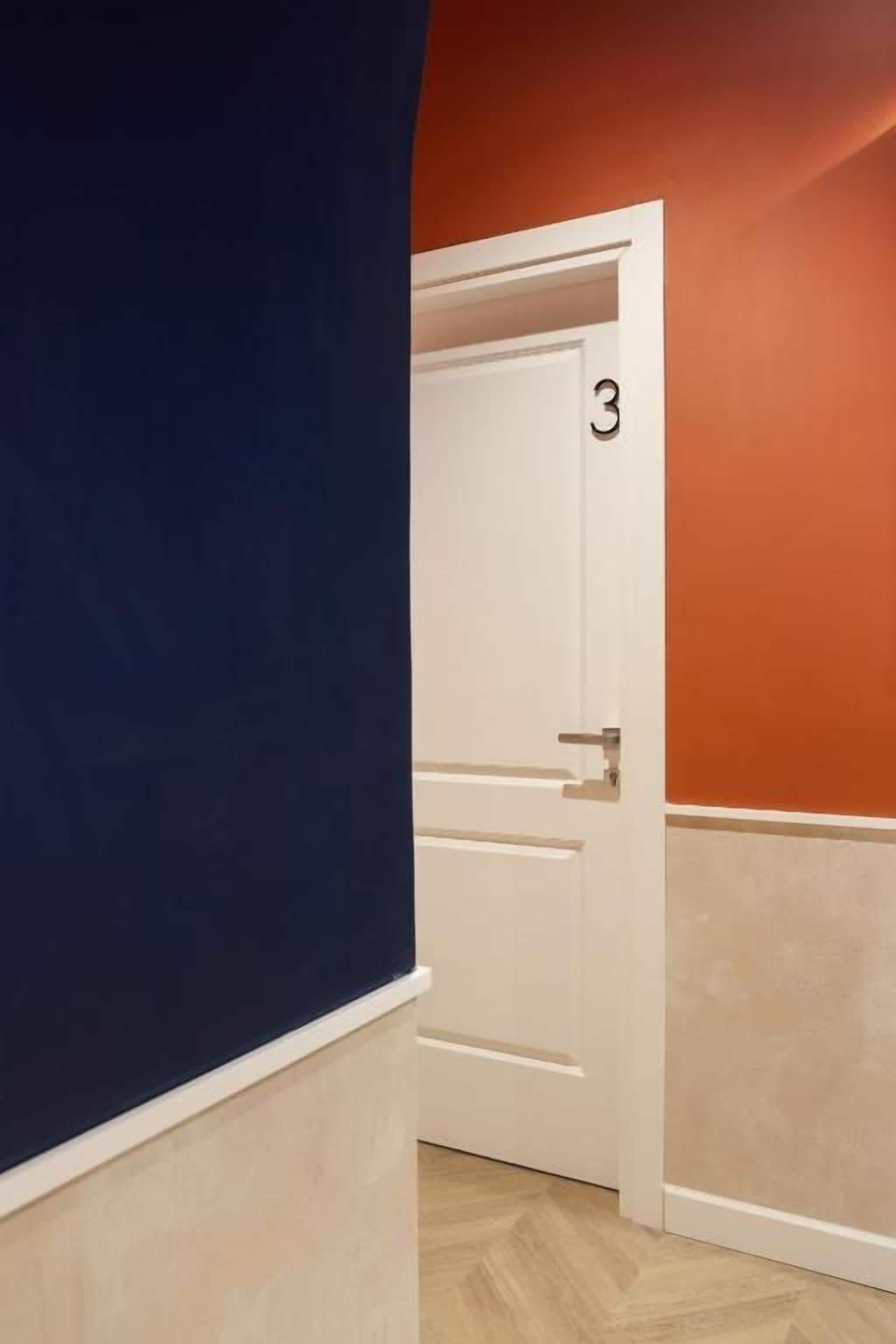 Un pasillo con una puerta blanca numerada con el número 3, suelo de madera y una vista parcial de una pared azul.