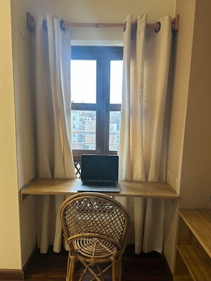 Ein Laptop auf einem Holzboden neben einem Fenster mit weißen Vorhängen, ein Korbstuhl davor und ein Blick auf Gebäude im Freien.