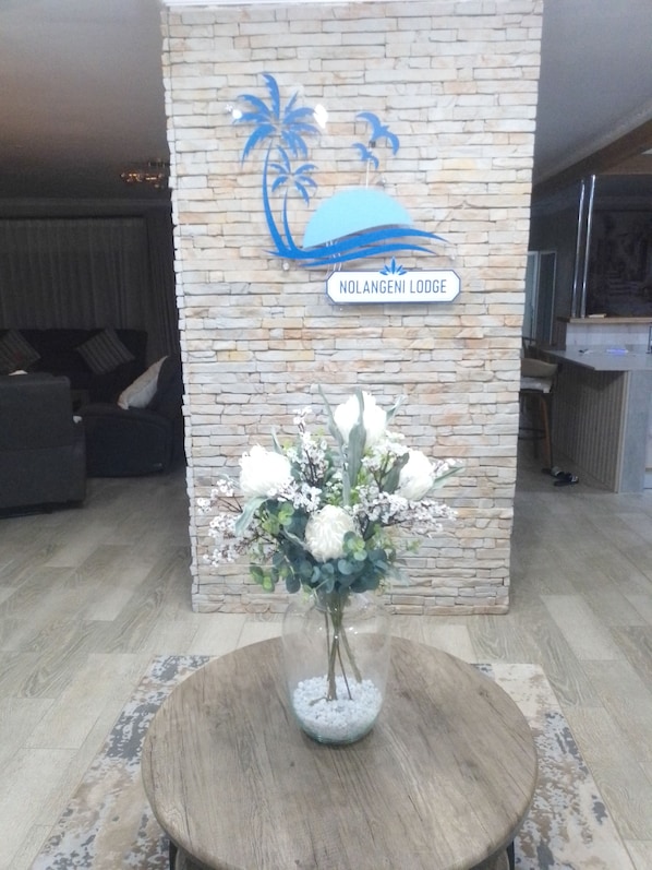 A reception area with a stone wall, a sign reading 'Nolangeni Lodge,' and a vase with white flowers on a wooden table.
