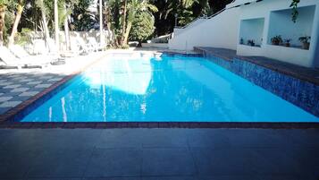 Une piscine dont le bord est recouvert de carreaux, entourée de plantes en pot et d’arbres.