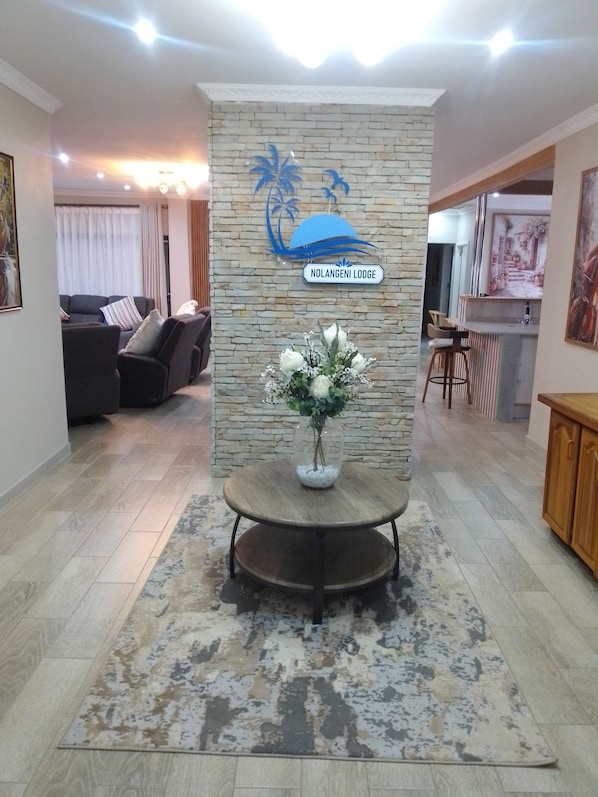 A reception area with a stone wall, a sign reading 'Nolangeni Lodge', a round table with a vase of flowers, and a seating area with sofas and a bar.