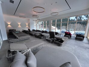 Ein moderner Wellnessbereich mit mehreren Liegen und Massagetischen, große Fenster bieten Blick ins Freie.
