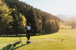 A person on a golf course with a golf bag and clubs, surrounded by trees and a clear sky.