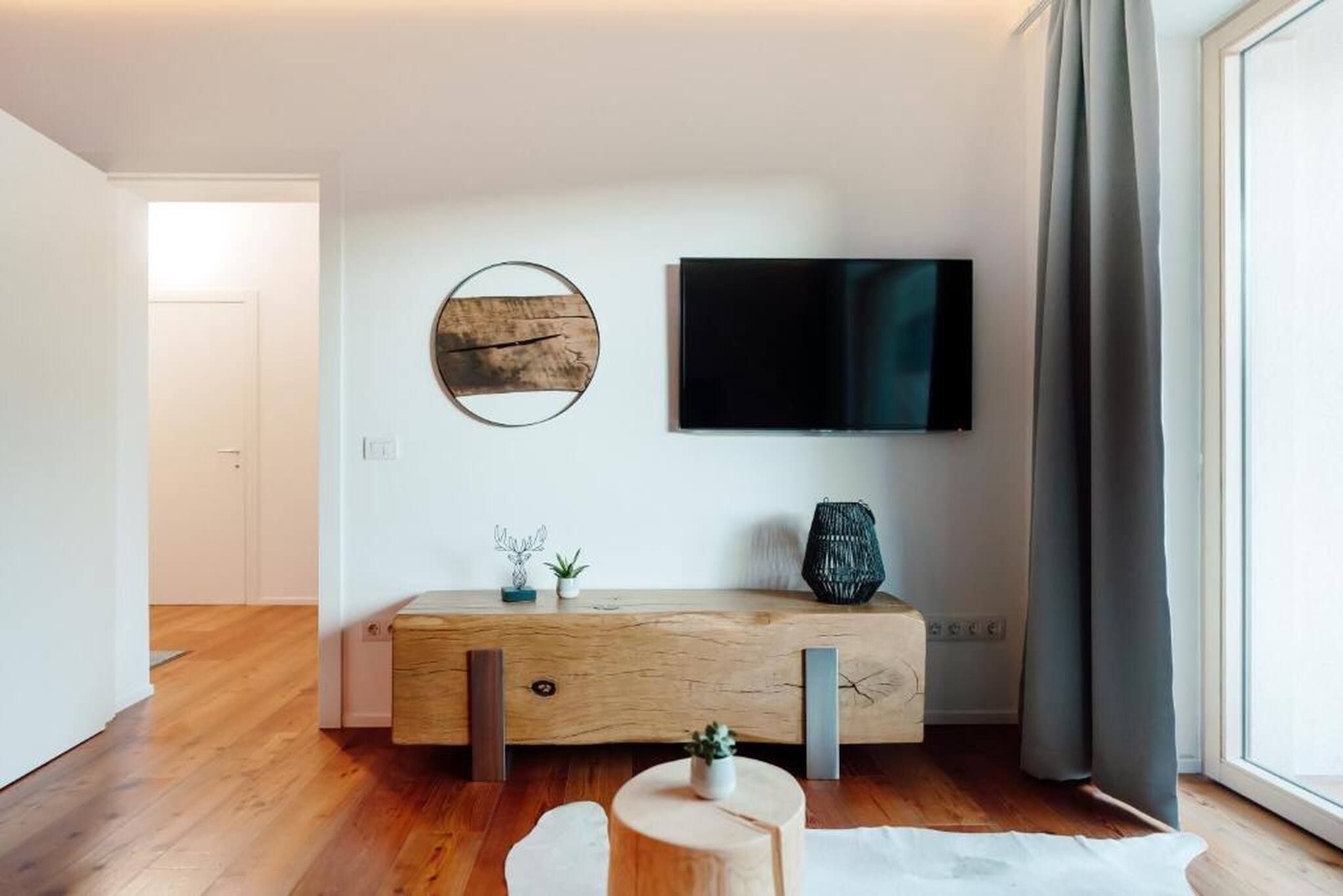 Een moderne woonkamer met een houten console, een flatscreen televisie aan de muur en een groot raam met gordijnen.