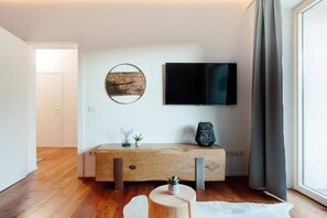 A modern living room with a wooden console table, a flat-screen TV mounted on the wall, and a large window with curtains.