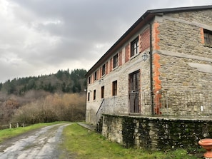 A stone building with a balcony, a dirt path, and a forested hillside in the background. - Casa Stabbi with Mountain View, Private Garden, and Air Conditioning (Chitignano)