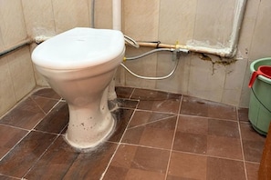 Un inodoro en un baño con piso de baldosas marrones y un cubo verde a la derecha.