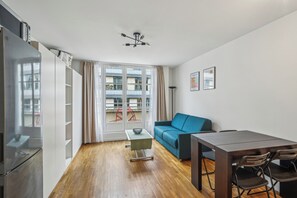 A modern living room with a wooden floor, a blue sofa, a dining table with chairs, a small coffee table, and a refrigerator. - ID 306 - Champs Elysées / Rue Euler (Paris)