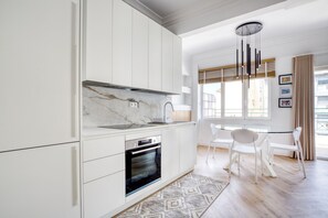 A modern kitchen with white cabinetry, a marble backsplash, and a built-in oven. There is a dining area with white chairs and a table, and a window with blinds. - Whitewood Residence (Athina)