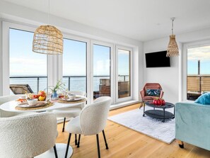 A modern living room with a large window, a dining area with a table set for two, and a seating area with a sofa and armchair. - Apartment in Olpenitz With Swimming Pool (Kappeln)