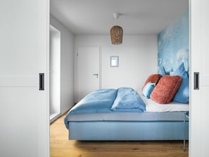A bedroom with a bed, a door, a wall with a painting, and a hanging light fixture. - Apartment in Olpenitz With Swimming Pool (Kappeln)