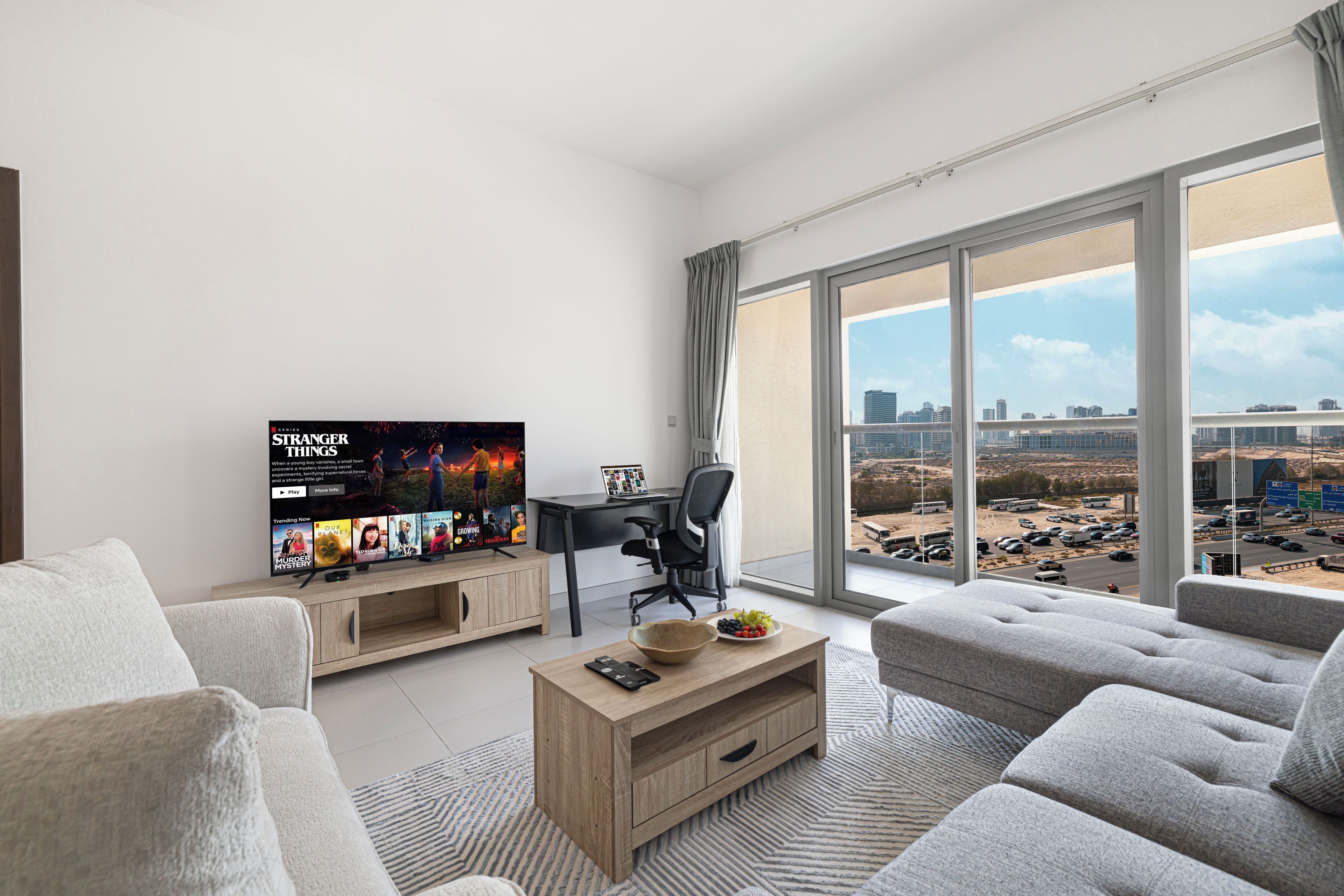 A modern living room with a large TV, a laptop on a desk, and a view of a cityscape through large windows.