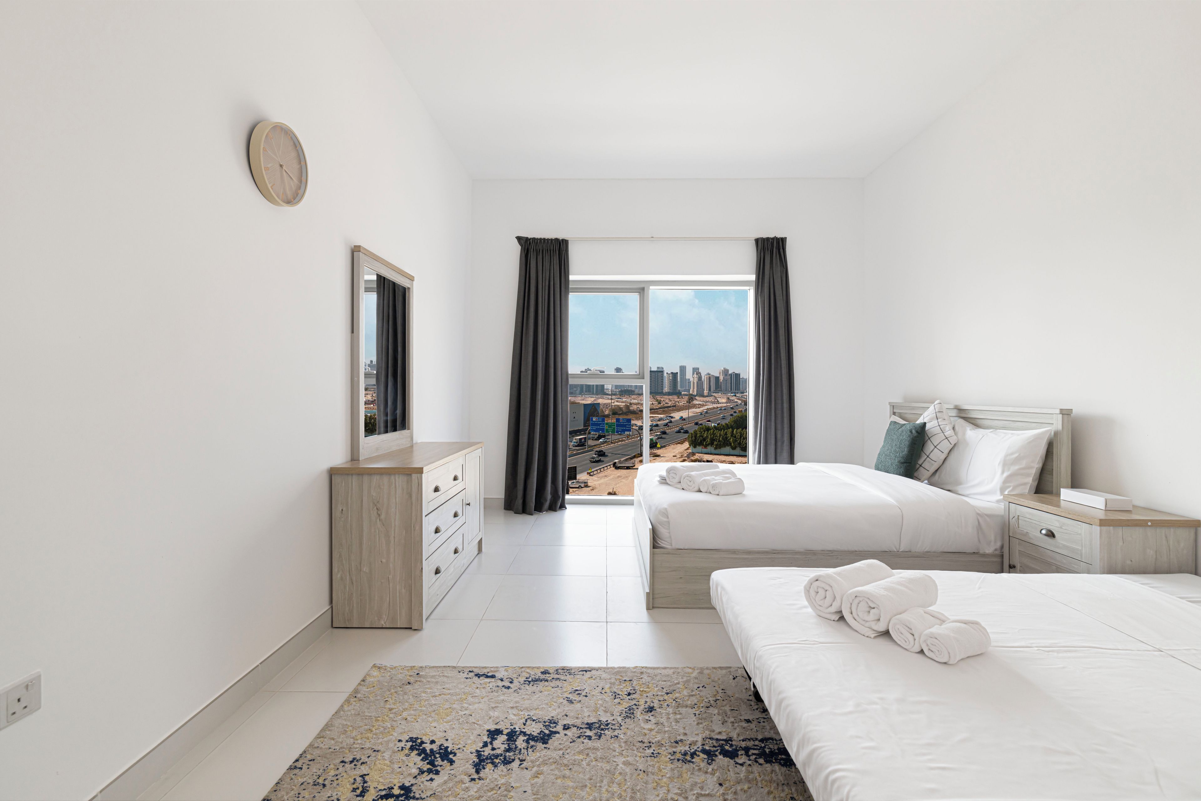 Ein modernes Hotelzimmer mit einem großen Bett, einem Nachttisch, einem Schminktisch, einem Fenster mit Blick auf die Stadt und einer Uhr an der Wand.