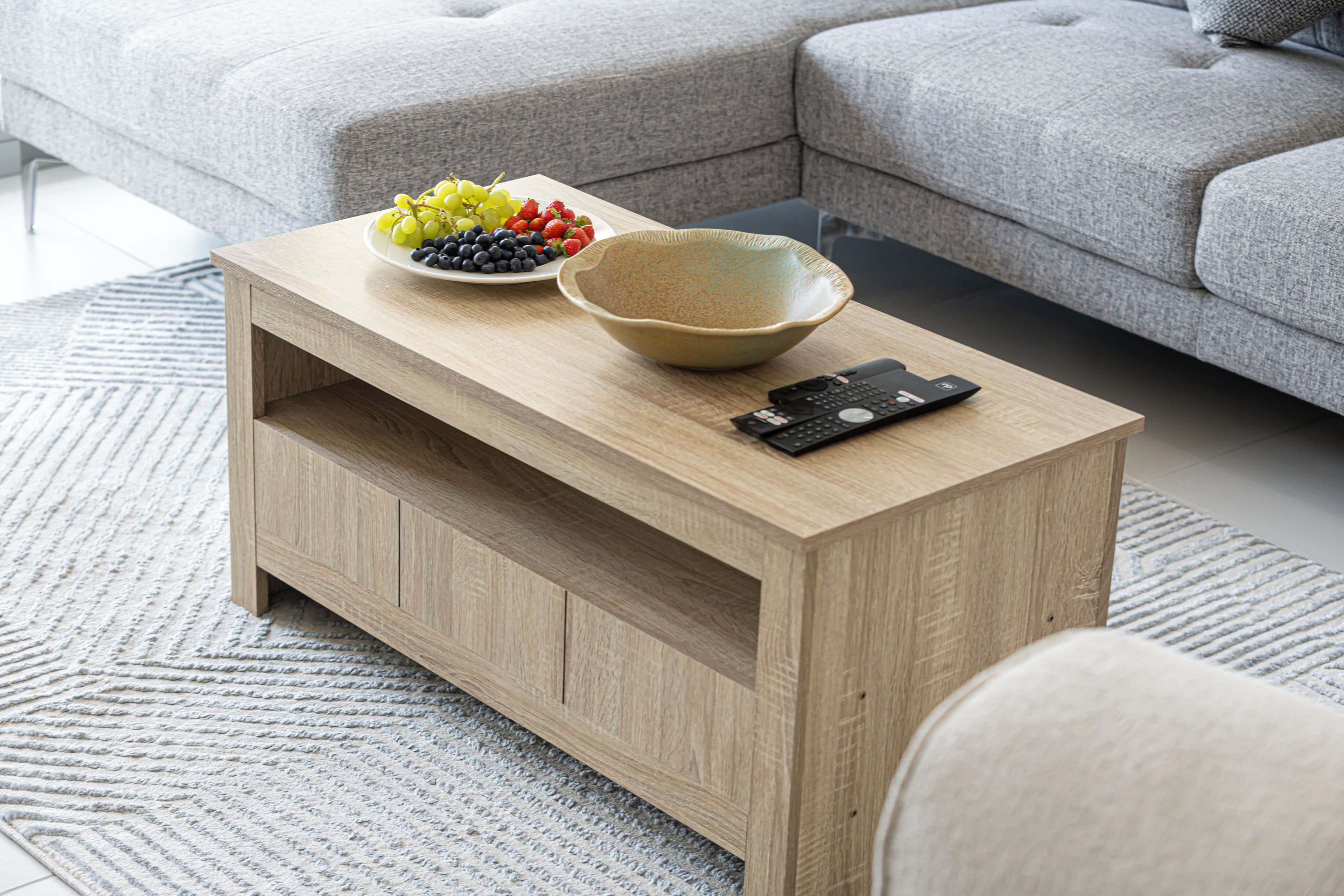 Ein Holztisch mit einer Schale und einer Fernbedienung darauf, ein graues Sofa im Hintergrund und ein gemustertes Teppich im Boden.