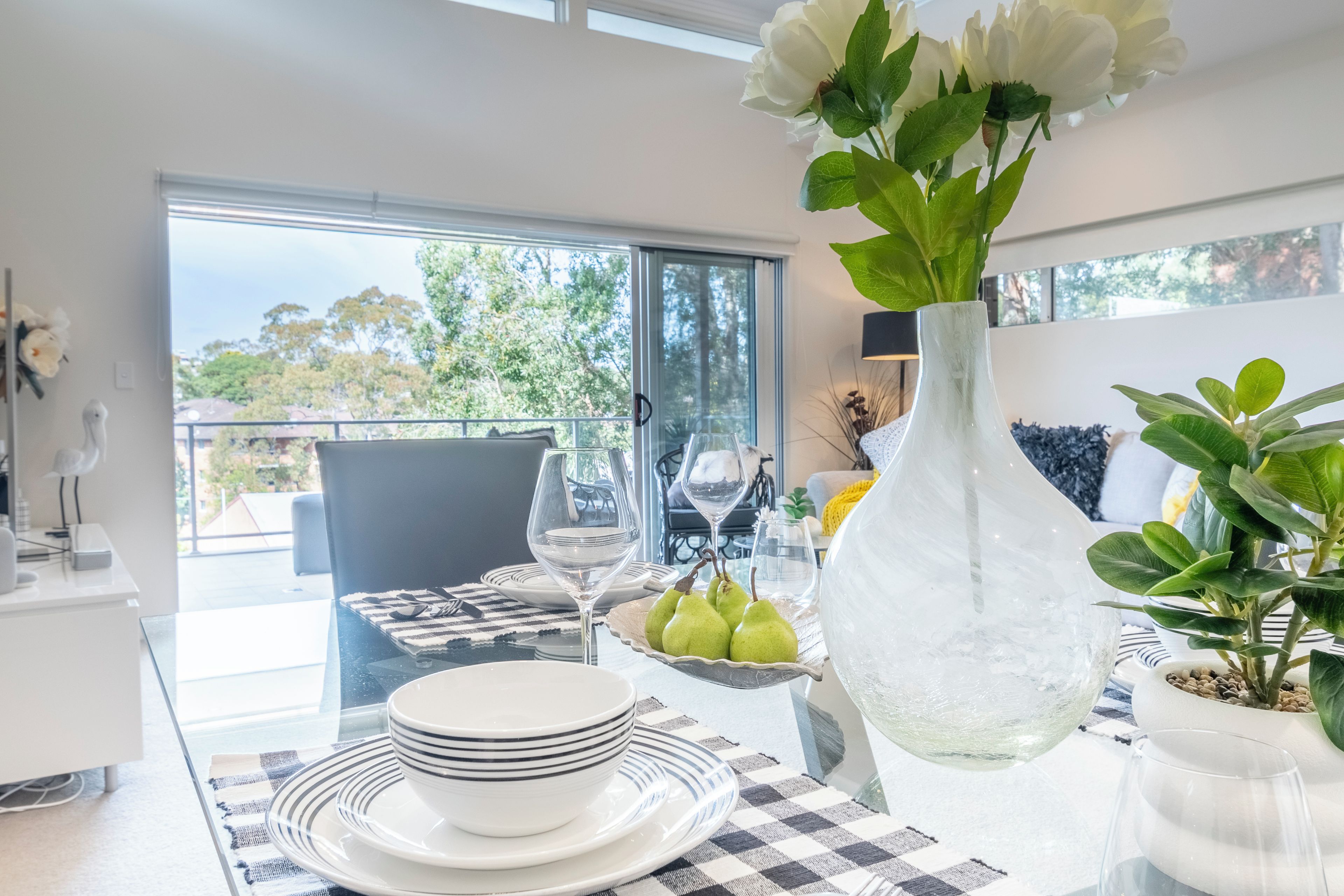 Un coin repas avec une table dressée pour deux personnes, un vase rempli de fleurs et une vue sur l’extérieur.