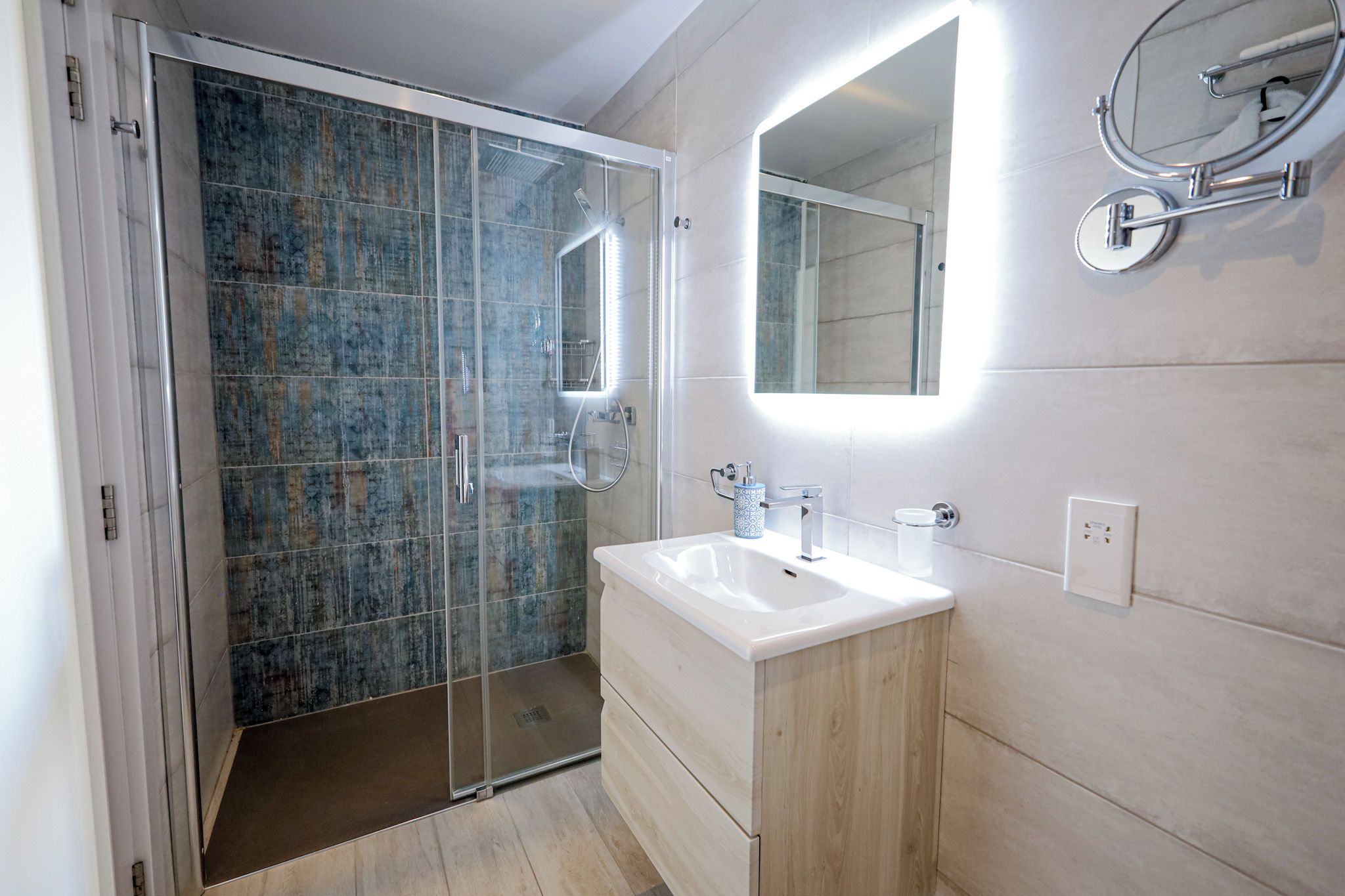 Un baño moderno con ducha envuelta en vidrio, un lavamanos blanco y un espejo con marco redondo.