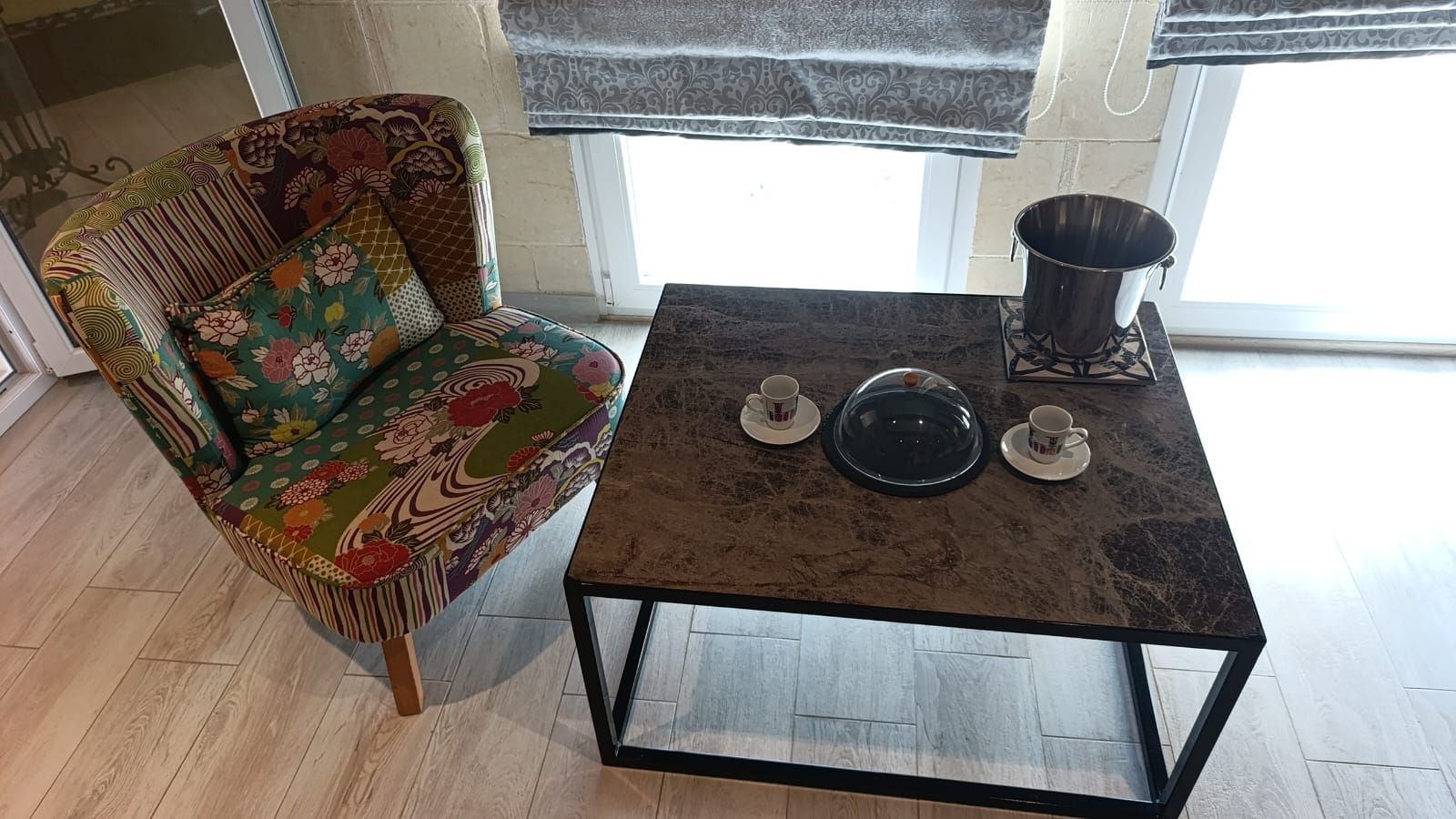 A room with a patterned armchair, a coffee table with a metal base, and a window with a curtain.