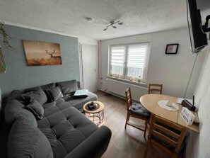 Una sala de estar con un sofá, una mesita redonda pequeña y una mesa de comedor con sillas. Hay una ventana con persianas, un radiador y un cuadro en la pared.
