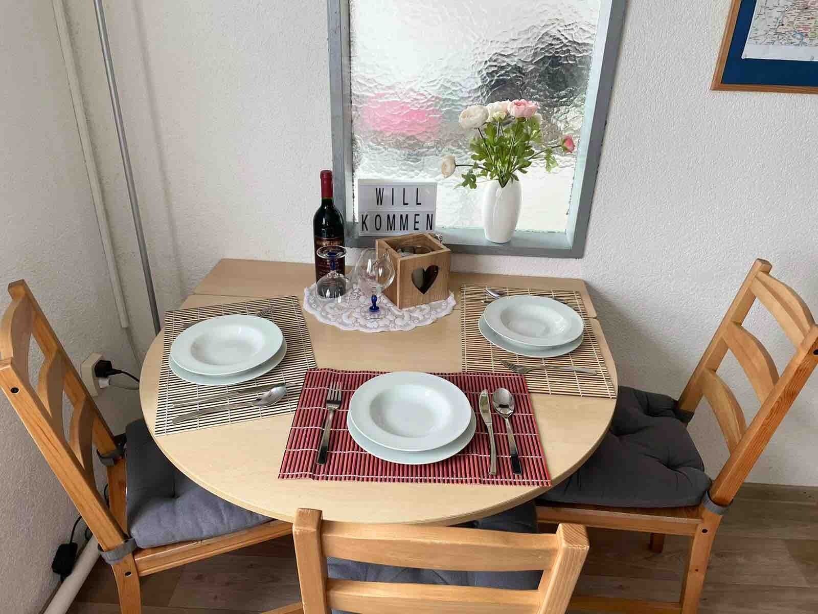Pyöreä ruokapöytä, jossa on kaksi tuolia, viinipullo, lasi, puinen laatikko ja kyltti, jossa lukee: ”WILL KOMMEN”.