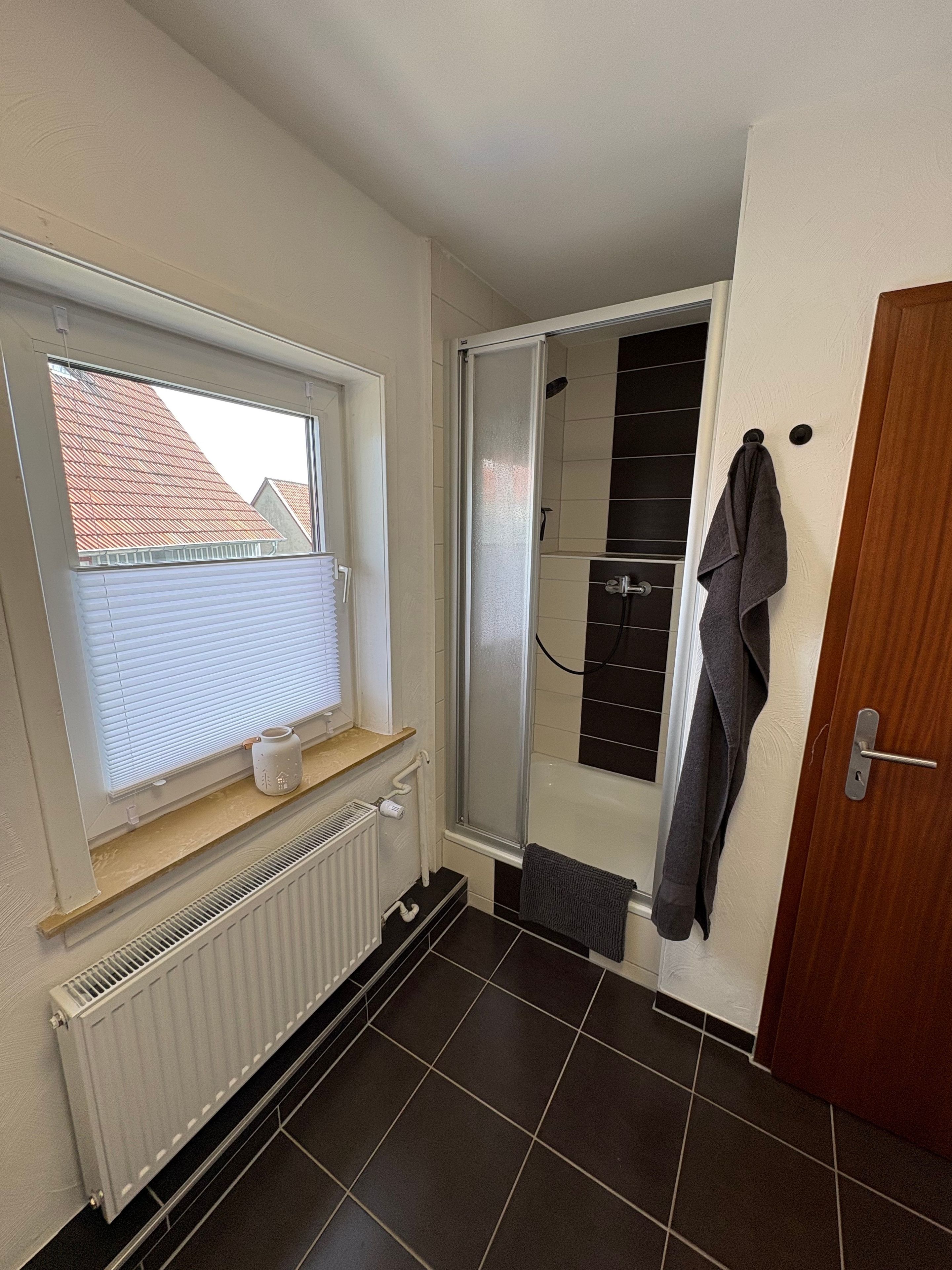 Ein kleines Badezimmer mit Dusche, Heizkörper, Fenster mit Jalousien und Handtuchhalter.