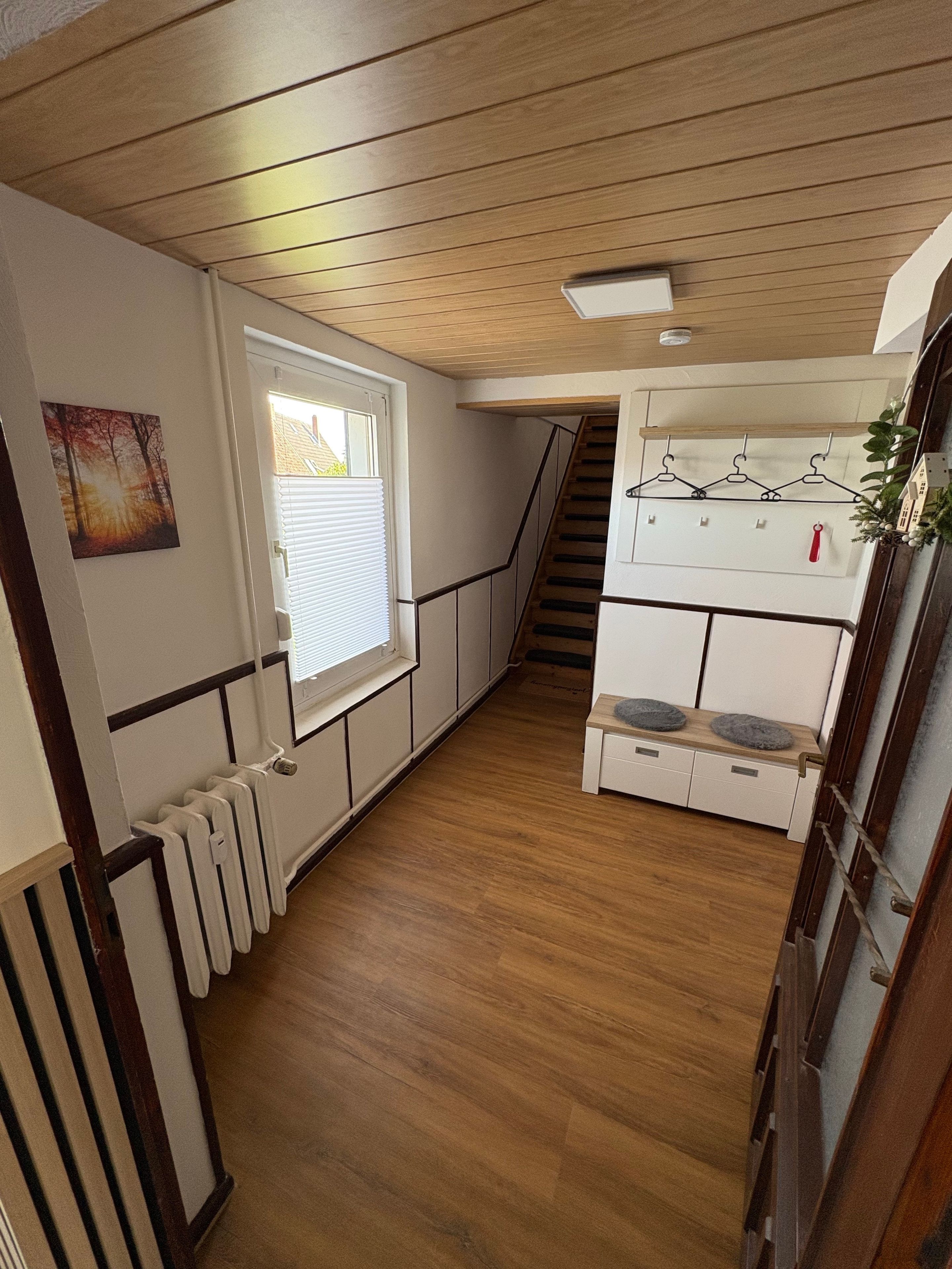 Un pasillo estrecho con suelo de madera, un radiador, una ventana con persianas, una escalera y una pequeña zona de kitchenette con un tendedero.