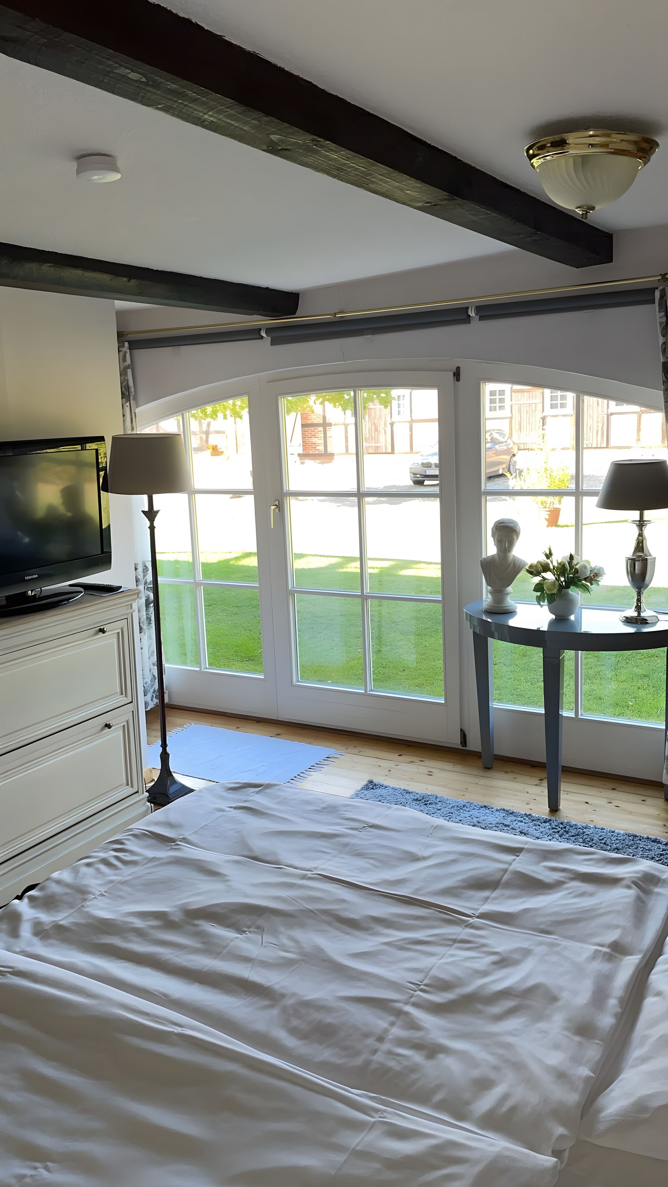 Ein Schlafzimmer mit einem Bett, einem Fernseher, einer Lampe, einer Büstenstatue und Blick auf eine grüne Wiese durch große Fenster.