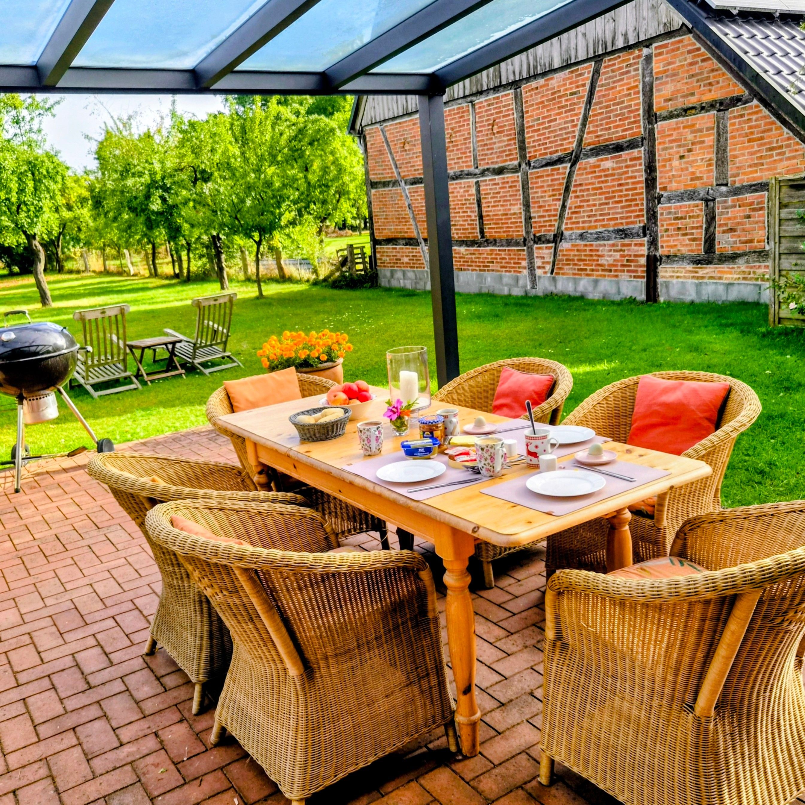 Un patio al aire libre con una mesa lista para comer, rodeado de sillas de mimbre y un muro de ladrillo al fondo.
