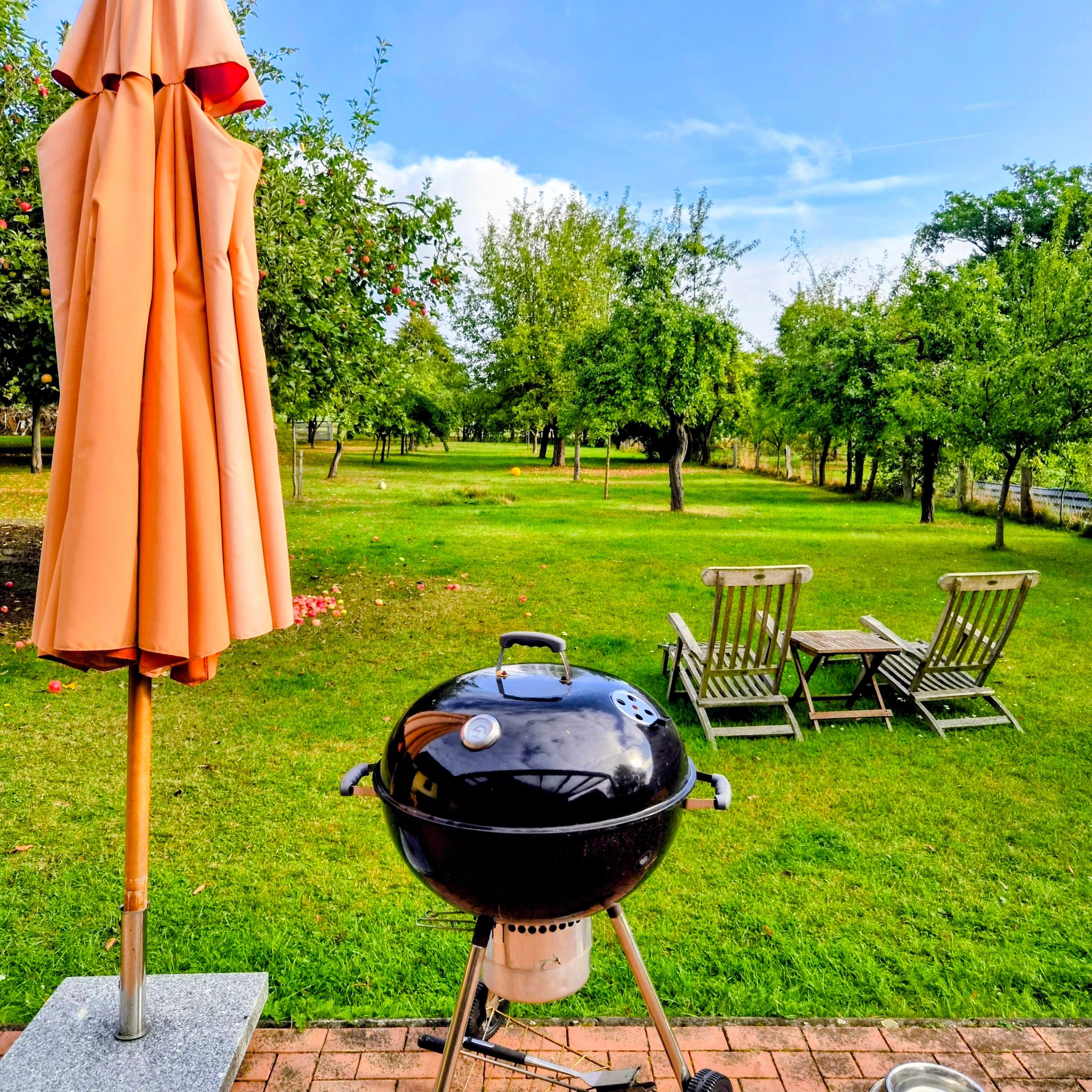 A barbecue grill in a grassy outdoor area with a patio umbrella and wooden chairs.