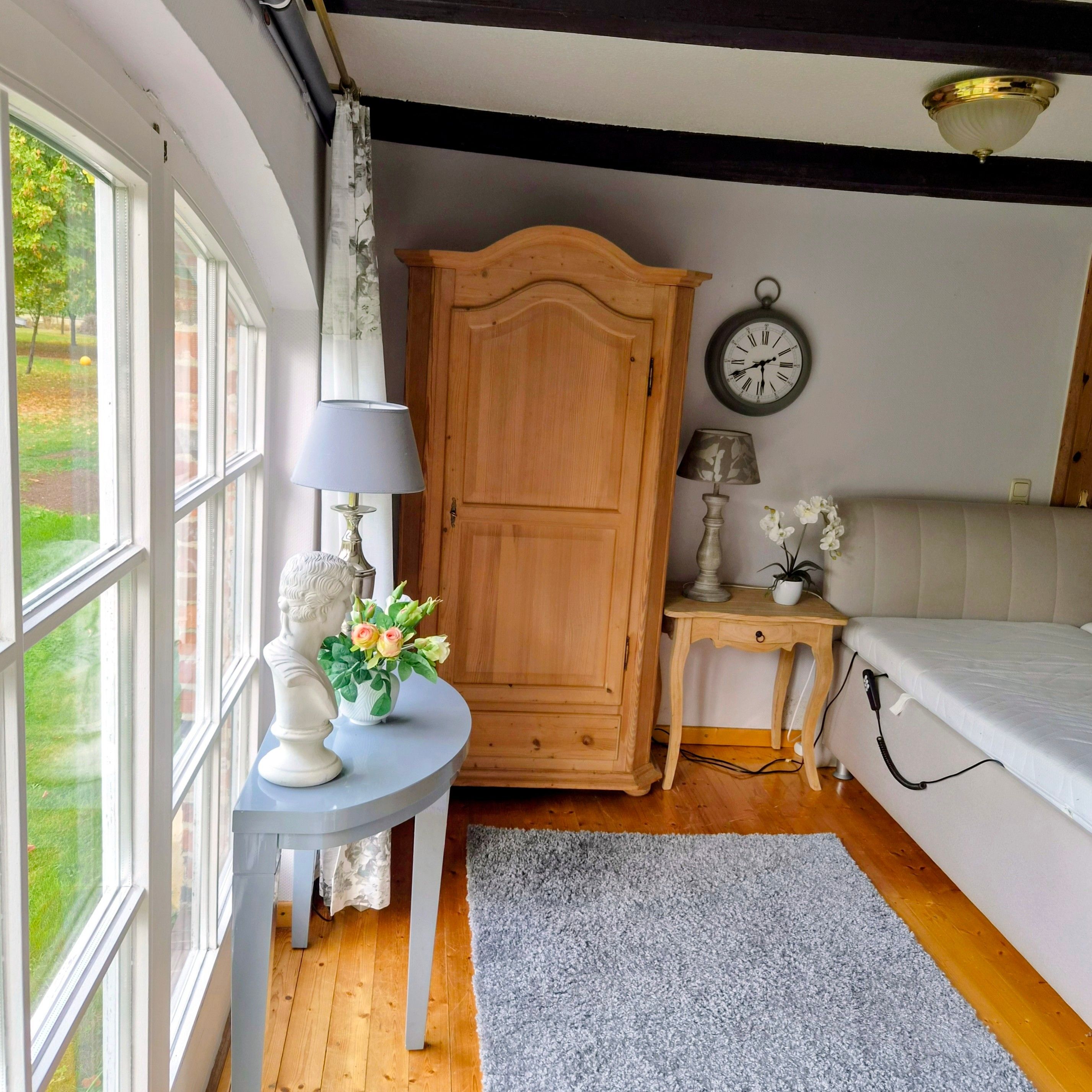 Una habitación con una cama, un armario de madera, un reloj de pared redondo, una mesita con una lámpara y un jarrón con flores.