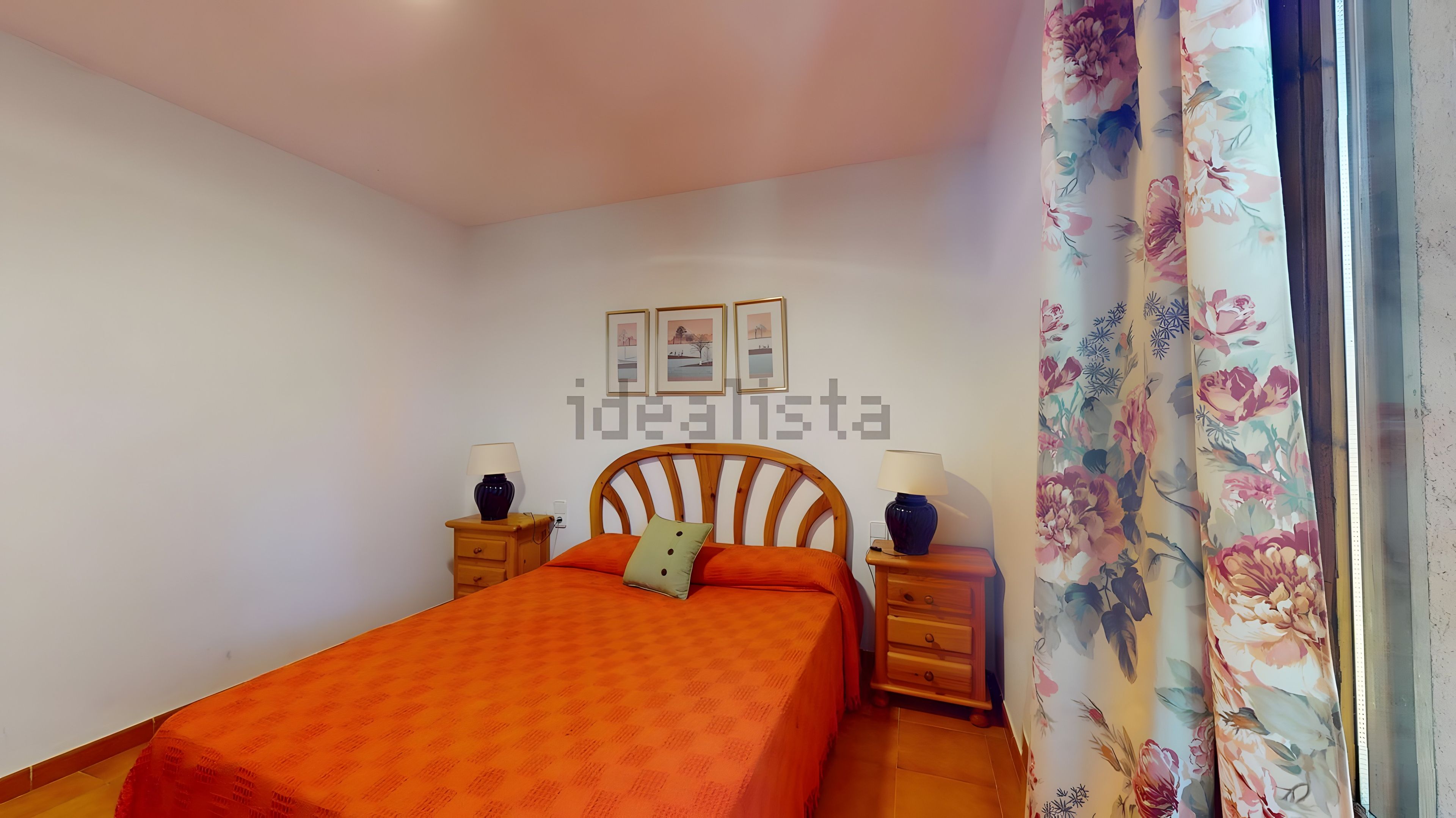 Une chambre à coucher comprenant un lit, deux tables de chevet, une commode, une fenêtre ornée de rideaux à motifs floraux et trois tableaux encadrés accrochés au mur.