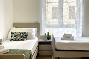 Een hotelkamer met twee bedden, een nachtkastje met een Boeddhabeeldje en een kamerplant, en een raam met jaloezieën.