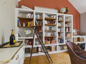 A room with a white bookshelf filled with books, a ladder, a wicker chair, and a wine bottle on a table. - House for 6, pool & garden, walk everywhere (Noirmoutier-en-l´Ile)