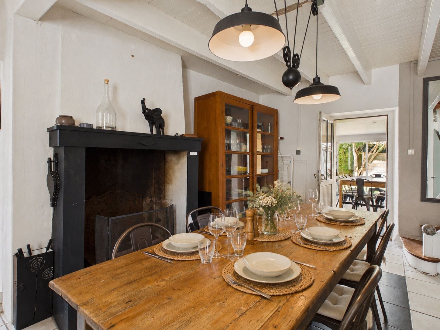 Un coin repas avec une table en bois dressée pour six personnes, une cheminée et des lampes suspendues.