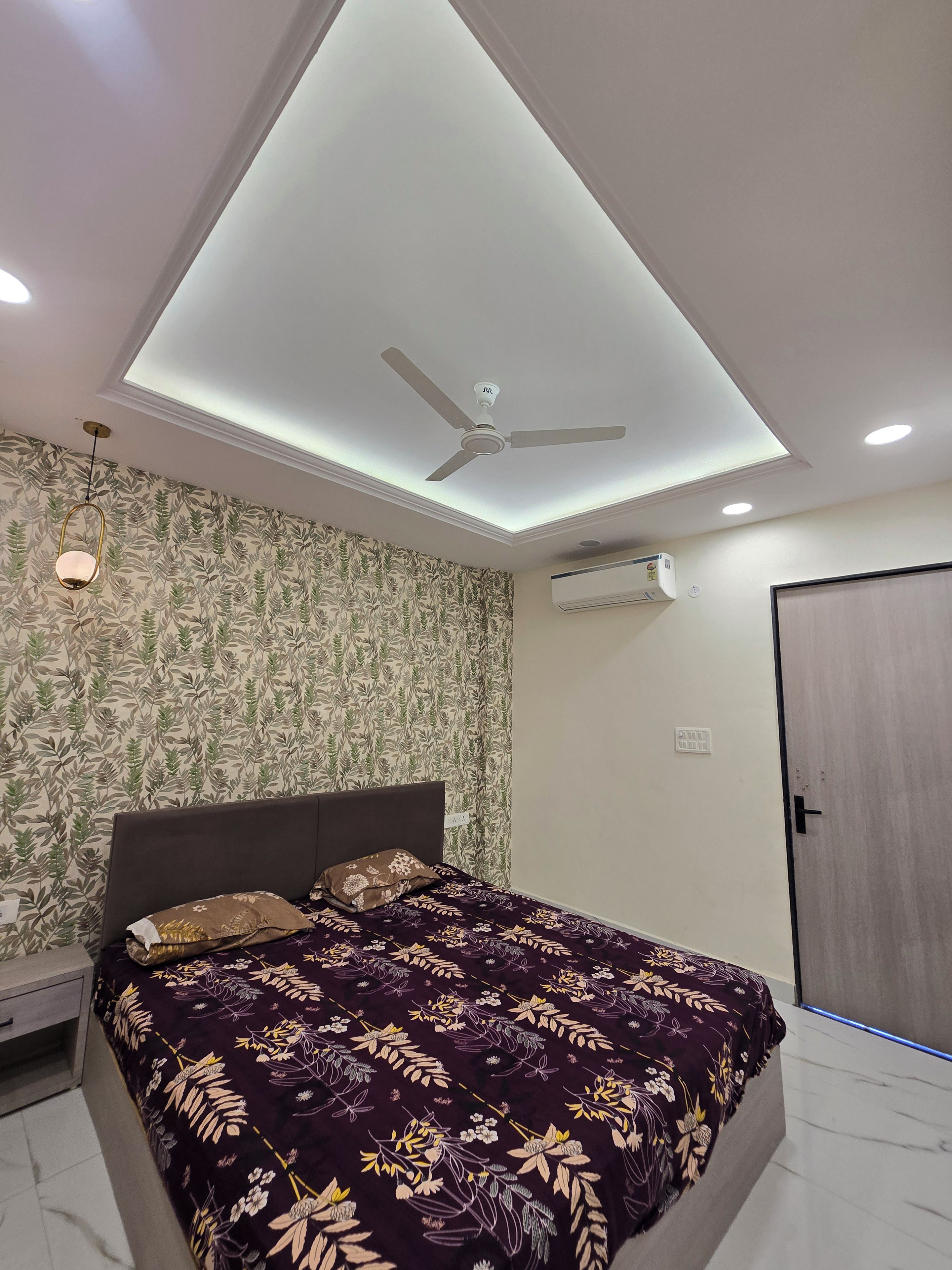 A bedroom with a bed, a ceiling fan, recessed lighting, an air conditioning unit, and patterned wallpaper.