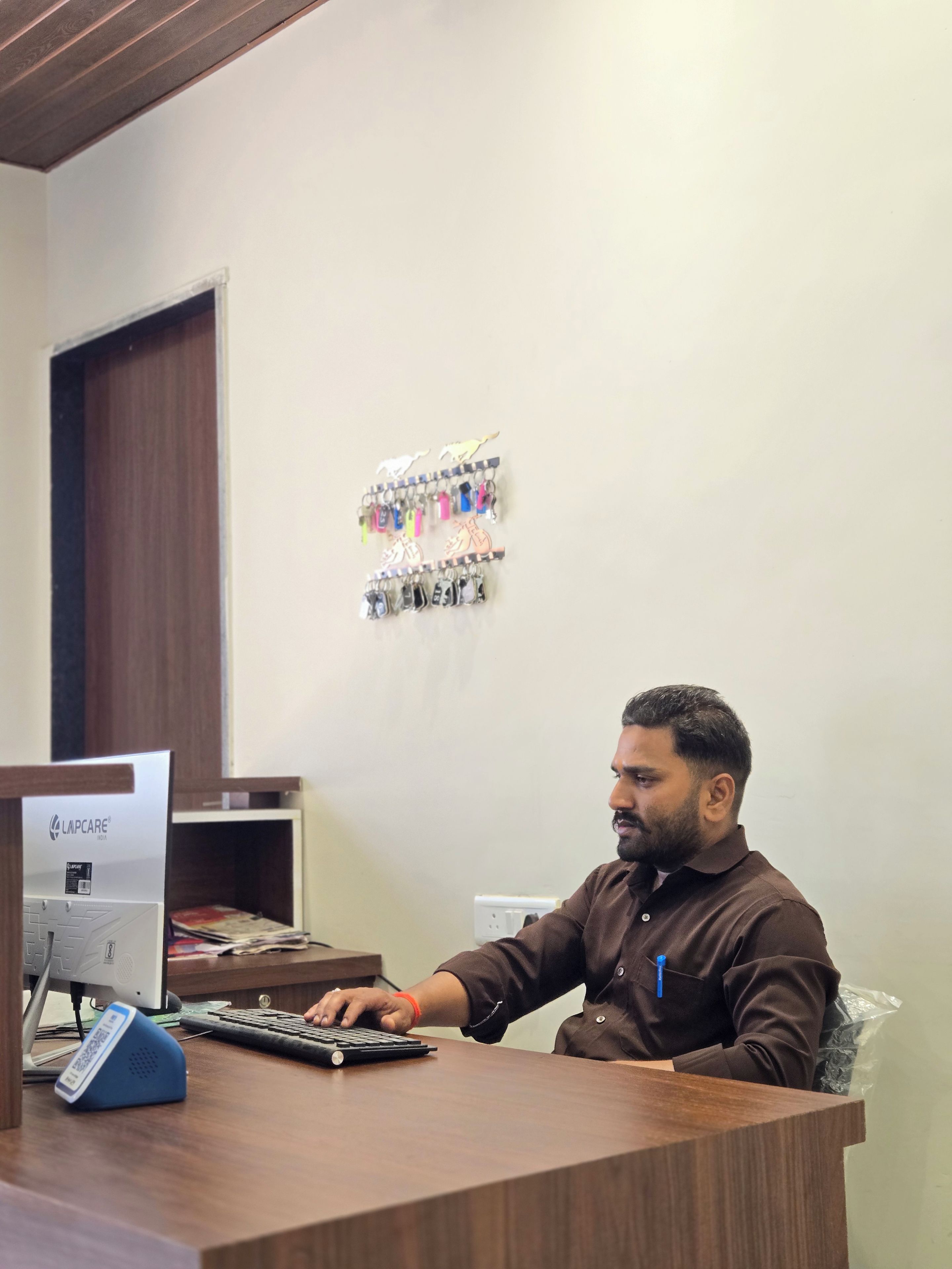 A person seated at a desk with a computer, a keyboard, and a mouse. There is a wall-mounted key rack above the desk.
