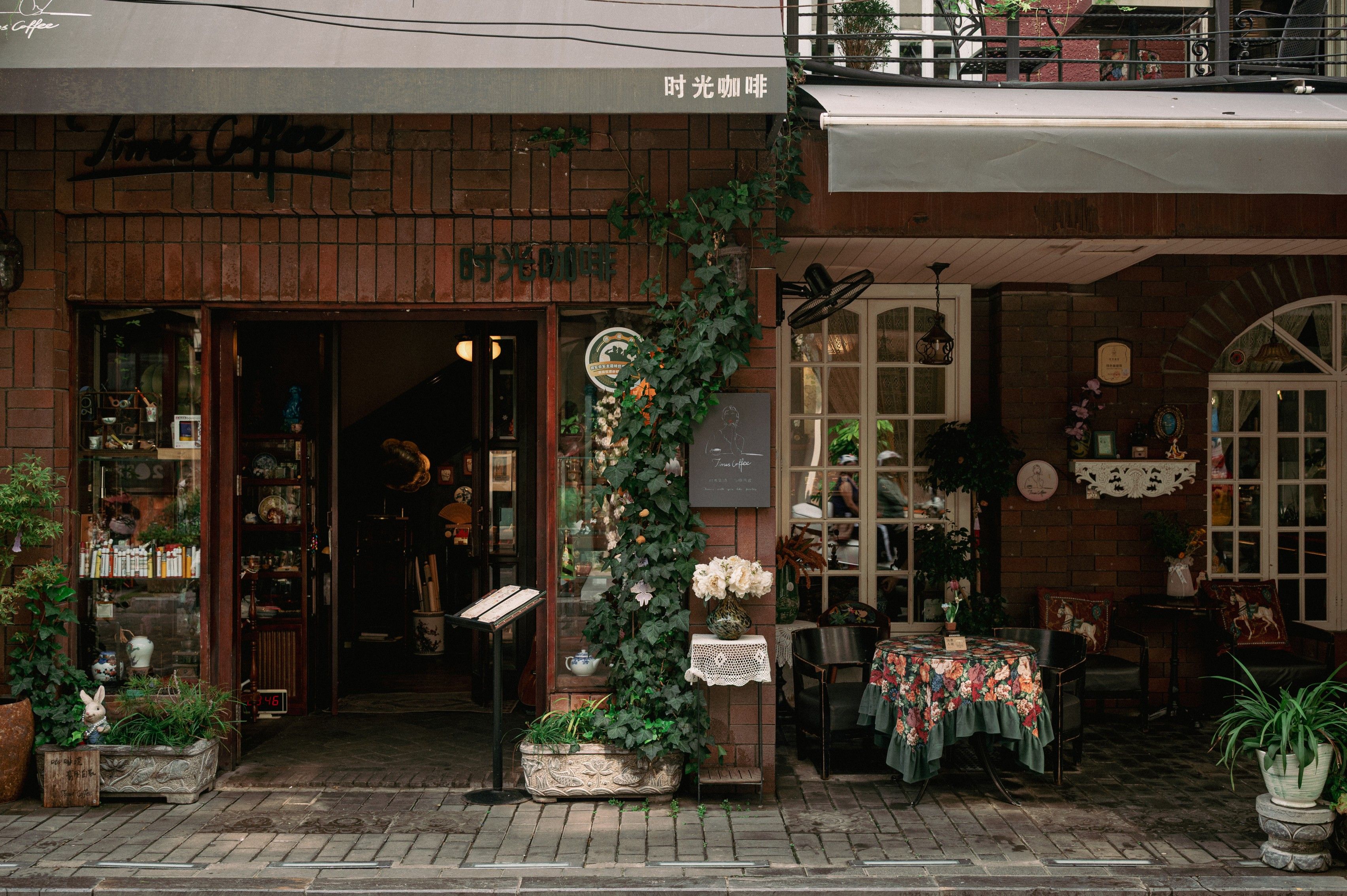 A coffee shop with a brick facade, outdoor seating, and a variety of plants and flowers.