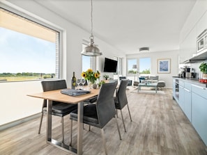 Eine moderne Küche mit einem Flachbildfernseher, ein Essbereich mit einem Holztisch und Stühlen sowie ein großes Fenster mit Blick ins Freie.