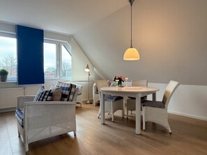 A modern living room with a dining area, featuring wicker furniture, a round table, and a large window with a view. - Holiday Apartment With 1 Bedroom (Wyk auf Foehr)