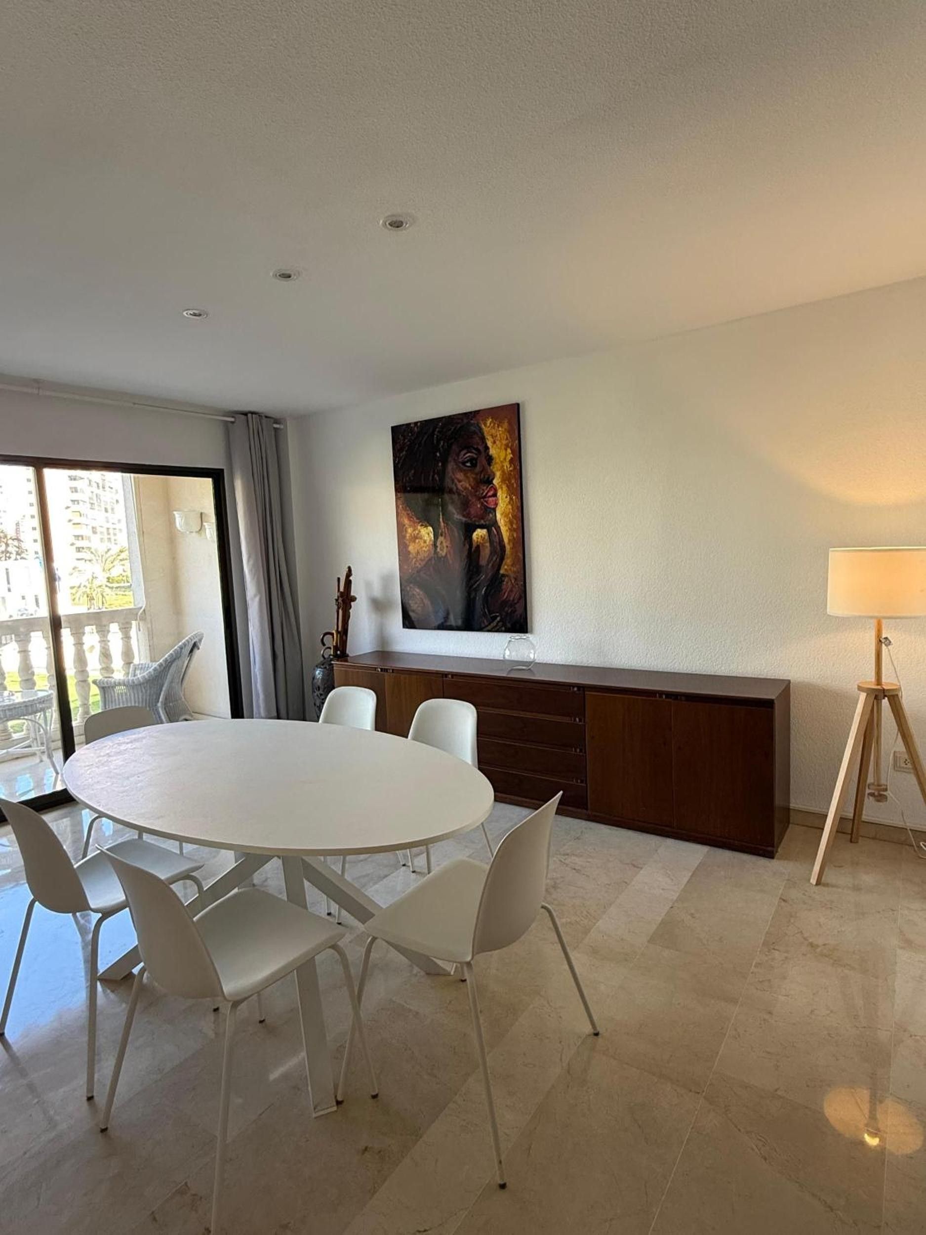 A modern dining area with a round table and chairs, a painting on the wall, and a lamp on a tripod.